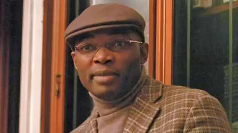 Charles Onana photographed in France in 2006 wearing a brown cap, a brown check jacket and beige turtleneck jumper