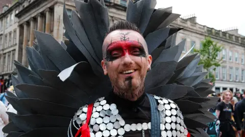 Pride parade attendee wearing large black wings, a silver circular mesh top and vivid red and black eye make-up smiles at the camera, standing on a Glasgow city street among other Pride attendees.