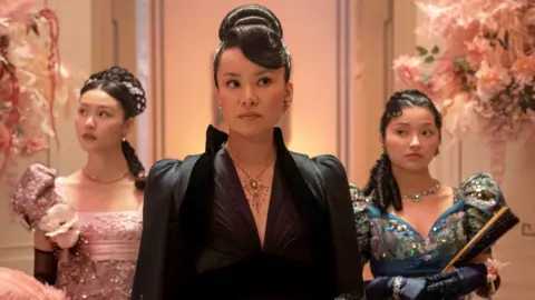 Araminta Gun stands in the centre of a pink floral hall entrance wearing a wavey hair updo, black dress and silver necklace. She is between her two daughters, one of whom is in a pink sparkling dress, the second is in a blue sparkling puff sleeve dress and wears navy blue gloves