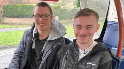 This picture shows best friends Tyler and Alfie on board a bus to York city centre. They are sitting next to each other on the lower deck, towards the back of the bus. Tyler is wearing glasses and a grey coat. Alfie is wearing a black hoodie and a white T-shirt. They are both smiling.