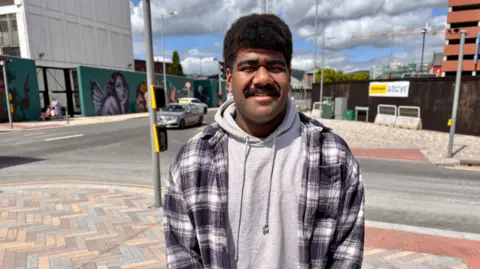 Livai Naga. He has think black hair and moustache, wearing a check shacket with grey hoodie. 