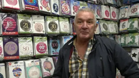 A man with grey hair, wearing a chequered shirt and dark coloured jacket is stood with a wall of age-themed birthday balloons in packets behind him.