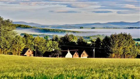 David Keir A green field stretches across the foreground, leading to a small cluster of houses and trees. Layers of woodland and open countryside extend into the distance, with a band of low mist hanging over the landscape. There is soft sunlight.