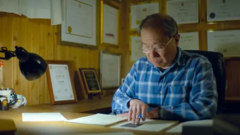Netflix Dr Shoo Lee, wearing a checked blue shirt, leans over a desk while holding a pen. He is looking over some documents, illuminated by light from a torch.