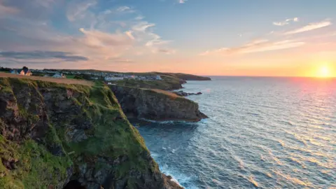 Clifftop view of Port Gaverne, green cliffs can be seen on the left of the image. The ocean is on the right. The sun is setting on the horizon.