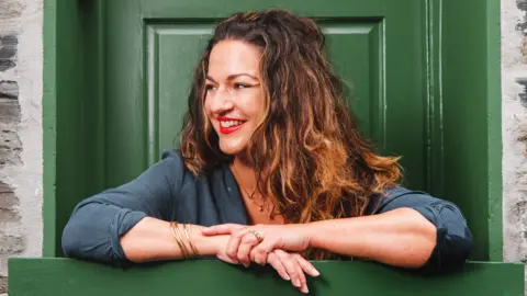 James Ram The picture shows Kate Attlee sitting in front of a green door. She has long, wavy hair with a mix of dark brown and lighter, golden highlights. The individual is wearing a dark shirt with sleeves rolled up to the elbows. On the wrists, there are multiple thin bracelets, and the hands are adorned with rings. The background includes a stone wall.