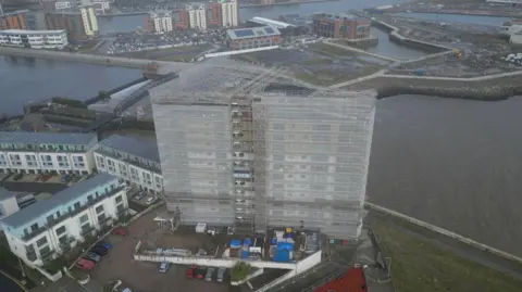 An aerial shot of a tower block covered in scaffolding with cars parked at its base. It is right by the seafront, with buildings and large patches of concrete on the other side of the water.