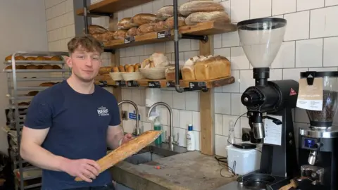 Toby Johnstone, gerente de harina, agua, sal, sosteniendo una barra de pan frente a estantes de pan en el café