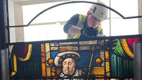 Holy Well Glass A large and colourful stained glass panel which shows Henry VIII handing a charter to Sir Thomas Vicary. The panel is full of rich yellows, blues and reds. Behind the panel is a man wearing a hard hat and high vis jacket, appearing to fix the top of the panel. It is unclear what tool he is using.