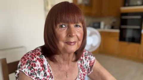 A woman with a red/brown bob wearing a white top with red and green flowers on it
