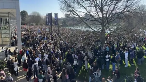 A very large group of people all standing outside on the UCD campus. The weather is bright and sunny. 
