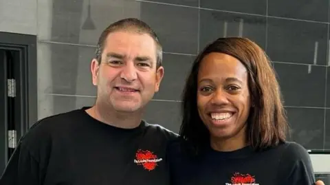 A man and a woman both wearing black T-shirts and black trousers look smiling at the camera with the backdrop of an empty cafe behind them. The man has has left arm around the woman's shoulder.