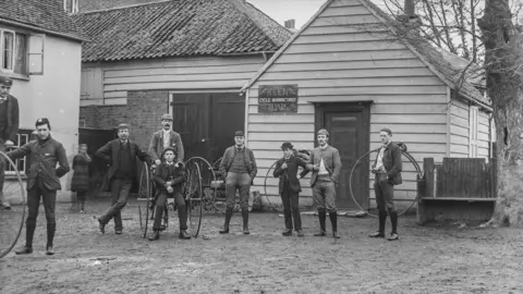 Historic England An historical photo of 10 people taken in Elmbridge. They are carrying large horse and carriage wheels, wearing boots and with hats.