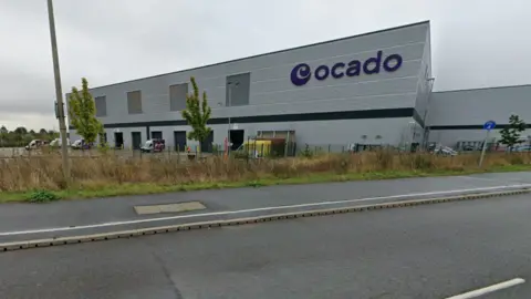 Google The exterior of the Ocado warehouse near Junction 11A of the M1 outside Luton. It is a grey building with a purple sign saying Ocado, with its logo.