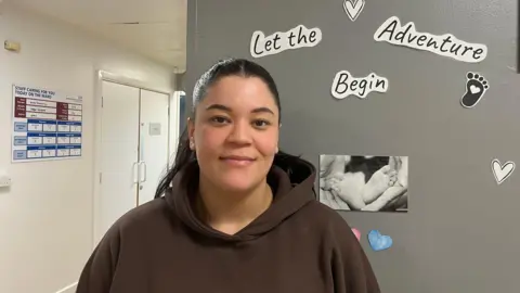 Jessica Bayley/BBC A woman with dark hair swept back in a ponytail wearing a brown hoodie is stood in front of a wall which has a sign that reads 'let the adventure begin' above a black and white picture of baby feet.