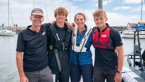 Melanie Dawes Harry Besley standing with Jacqui Besley, Henry Cietak, and another crew member, they all have their arms around each other and are standing in Lyme Regis harbour, with sailboats visible in the background. 