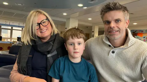 A young boy is sitting between his mum and dad. The boy has short brown hair and is wearing a blue t-shirt.  He is staring at the camera. His mum has dark framed glasses on and blonde short hair.  She is wearing a large scarf and smiling at the camera. The boy's dad is sitting on his left and is wearing a cream knitted jumper, he has grey spikey hair. 