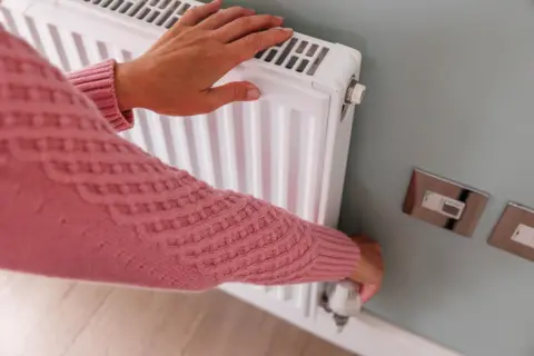 A woman wearing a pink woolly jumper is touching a radiator with her left hand while adjusting the knob on the radiator with her right hand, 