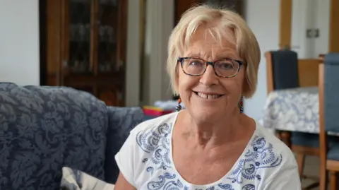 Shaun Whitmore/BBC Jane Goose sitting down on a blue patterned sofa. She is looking directly at the camera and smiling. She is wearing a white short-sleeved T-shirt with a blue pattern around the neck line and has a pair of glasses on and dangly earrings.