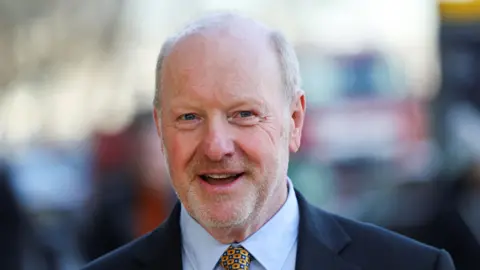 Former sub-postmaster Alan Bates smiling at the camera while wearing a dark suit and yellow tie. 