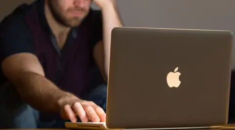 PA Media An anonymous man sitting in a dark room with his Apple laptop on a table in front of him. The laptop screen is lighting up his face and he has one hand resting on the mousepad and the other holding his temple. 