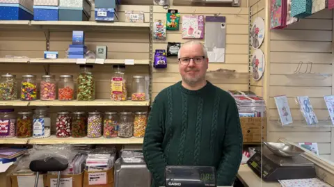 A man with short grey hair and a beard stands behind a till in his shop. He is wearing a thick green jumper and black-rimmed glasses. Jars of various sweets stand on shelves behind him. Cards, boxes and other items can be seen. There is a set of scales.