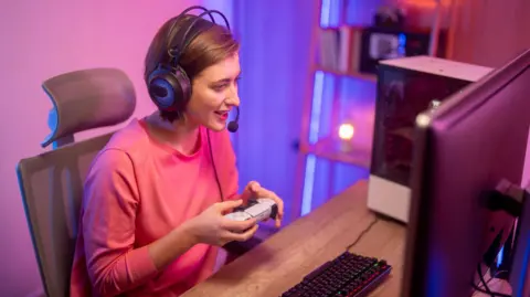 A female streamer is sitting down at a desk looking at her computer, holding a white and black PlayStation 5 controller. She is wearing a pink jumper and has a black headset on.