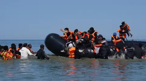 Migrants climb onto small boat in sea that is around waist-height. They wear orange life jackets