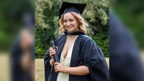 NHS Humber Health Partnership A portrait photo of a woman with shoulder length blonde hair which has moved back a bit in the wind. She is smiling and wearing a low light-yellow dress with a gold necklace and a black graduation gown, cap and rolled up certificate in a box.