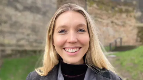 Nottingham City Councillor Sam Lux is pictured smiling outside Nottingham Castle. She has long blond hair and is wearing a grey coat. 