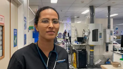 A woman wearing round, gold-rimmed glasses and hair scraped back into a ponytail with a dark blue medical tunic with hospital machines in the background.