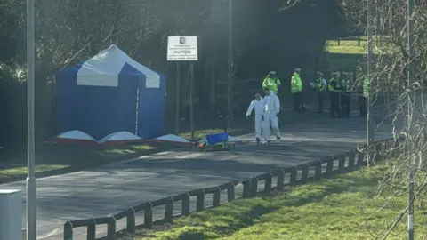 Handout Two forensic investigators in white PPE stand on a road. A blue tent has been erected on a path on the pavement and a group of police officers stand in the background.