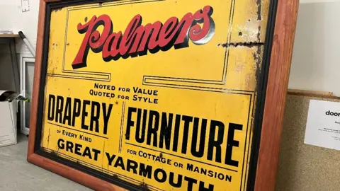 Andrew Turner/BBC A yellow sign with a wooden frame. On the sign if red writing which says Palmers. There is also black writing on the sign which says Drapery and Furniture.