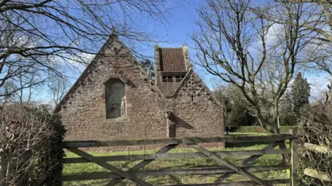 A village church surrounded by daffodils in the sunshine. A five bar gate is in the foreground.