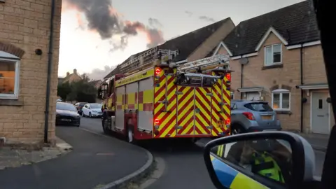Malmesbury Police A fire engine navigating through the narrow roads of a housing estate with cars parked on either side of the road. Smoke can be seen in the distance. A wing mirror of a police car is visible on the lower right, with a police officer seen taking the photo.