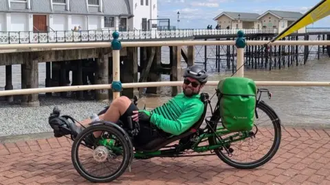 Rhys Holmes A man in a modified green bike. He's wearing a black helmet, green cycling too and black shorts while being sat down in his Recumbent Cycle. 