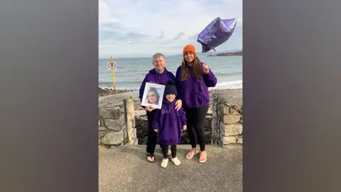 AMFF Sian Ashcroft’s mum, Sue Dowling, sister Lauren Dowling and Lauren’s son stand in front of a wall above the sea on Traeth Bychan beach in Anglesey. They wear purple jumpers; The mum holds a framed picture of her daughter. Lauren Dowling holds a purple star-shaped balloon.