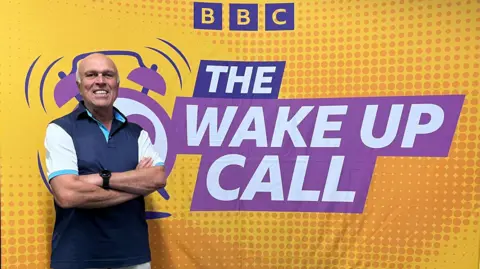 A picture of Steve Holt stood in front of 'The Wake Up Call' sign on a yellow poster. He is wearing a white shirt and a navy jacket.