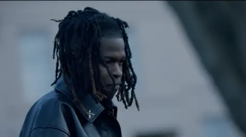 Lindsay Entertainment Jordan Adetunji. He has shoulder-length dark hair, wearing a black leather jacket. He is looking to his side.