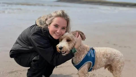 Family photo Emily is on a breach hugging a terrier type dog. She has long blonde hair swept back and is wearing a black windbreaker jacket and matching trousers. She is smiling and holding her cheek against the top of the dog's head. 