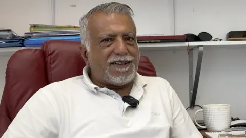 Shop owner Bipin Chawla, who is a middle aged Indian man, with grey hair and a grey short beard, sat in his shop office in a red leather computer chair, wearing a white polo shirt, with a white coffee mug showing on his desk next to him