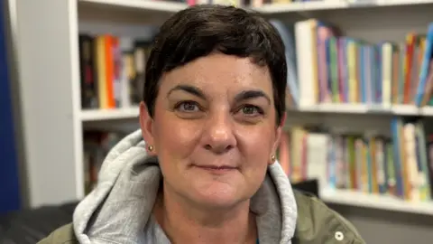 A woman with short black hair wearing a grey hoody and green jacket. There are bookshelves with books behind her.