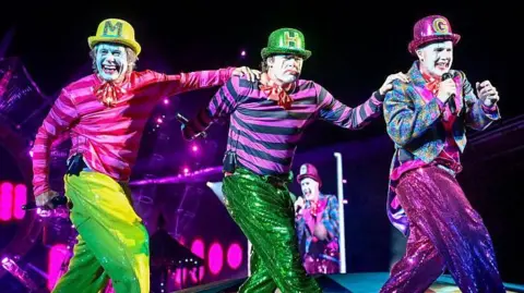 The three men in pink and green circus outfits and face paint performing on stage in a row. They are all wearing bowler hats with the first letter of their name on. Gary Barlow holds a microphone up to his mouth, while Mark has his hand on Howard's shoulder, and Howard has his hand on Gary's shoulder. They all wear sequin trousers. 