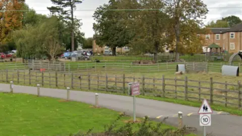 Google view of the Acacia Hall site. There is a long tarmacked driveway leading up to a large brick building, with cars parked outside.
