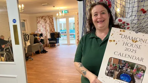 Allison Webster smiles at the camera as she holds up a calendar with "Diamond House 2025" written on the top. She is wearing a green polo shirt and has brown hair. A number of residents are sitting in a room behind her. 