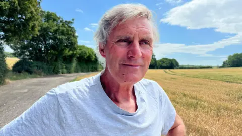 Farmer Will Dickinson is stood on a sunny day near a field of wheat.