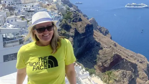 Family handout Ellen Mulvey on holiday abroad with long blond hair wearing black sunglasses and a white and blue sun hat and lime green t-shirt with the words 'The North Face' on it. She is standing on a property on a hill by the sea. She is smiling.