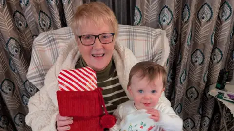 Grandmother Pat grant sitting on a chair smiling at camera holding her baby granddaughter and a Christmas stocking. They're both smiling. 