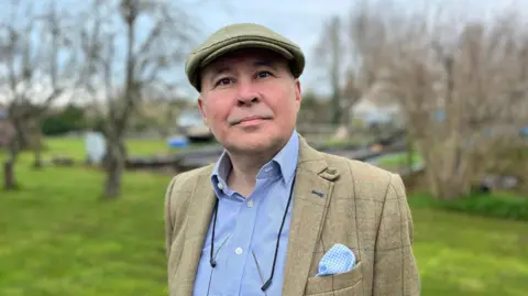 A man with a green flat cap looking towards the camera. He's wearing a blue shirt and a green jacket with a blue and white checkered pocket square. 