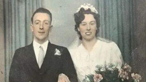 Image taken of Tessa Gay and her husband in their wedding outfits at a photograpy studio. He is on the left wearing a smart black suit and grey tie while she is in a white dress and wearing a floral headband and wedding veil behind her, holding a big bouquet of flowers.